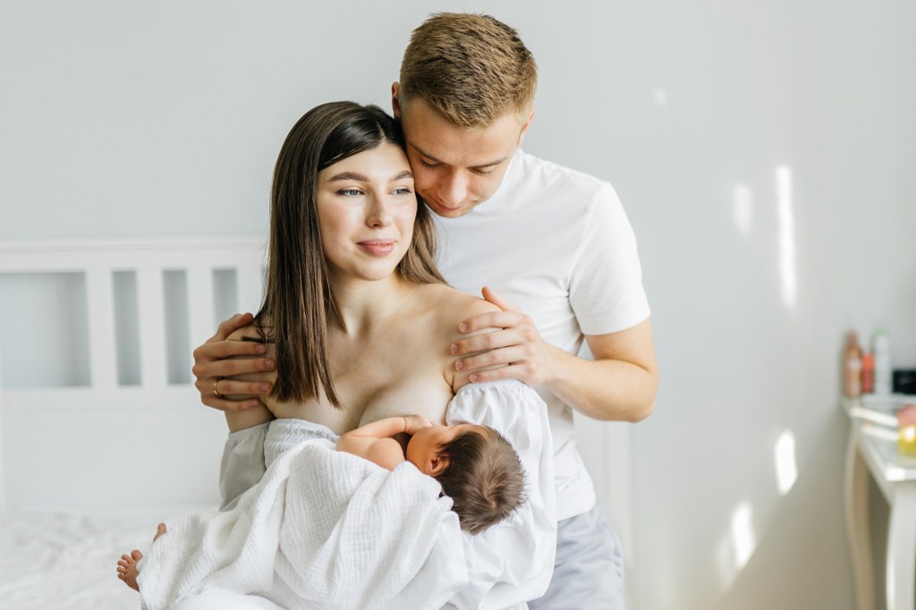 Couple in love with similar Mario Novembre, sitting in a cozy bedroom; the woman breastfeeding her newborn daughter while her husband embraces her warmly.