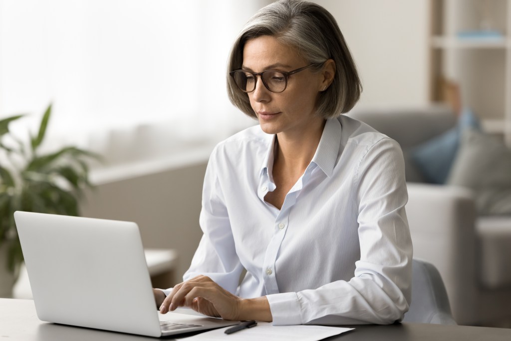 Focusing quite mature business woman in elegant cups that print on the laptop, working on the computer in the house office, using internet technology for independent work communication
